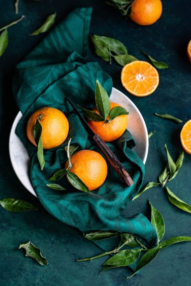 Fresh oranges arranged in a bowl with green leaves and a teal cloth backdrop.