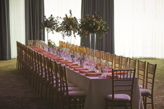 Elegant long dining table set with flowers and tableware, surrounded by chairs.