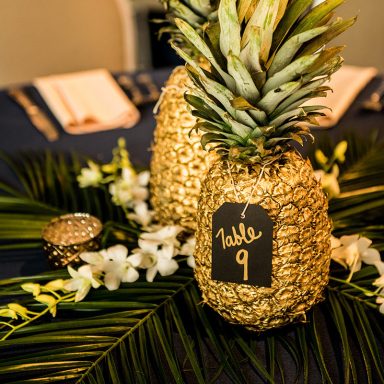 Gold pineapples with a table number and flowers on a dark tablecloth.
