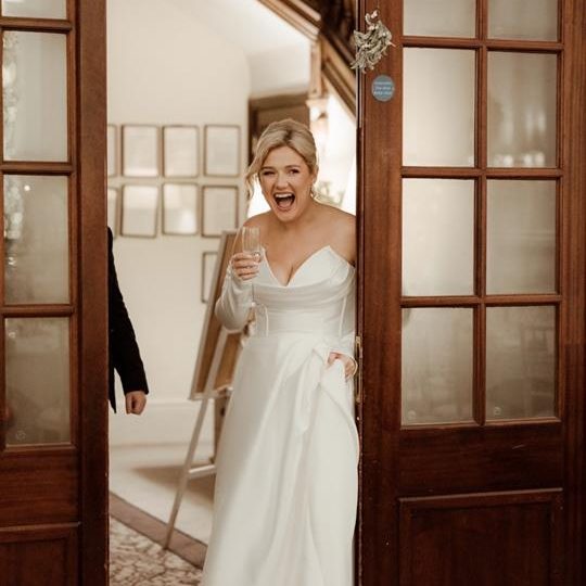 Bride in a white dress joyfully emerging through double doors, holding a glass. Seeing  venue  decor for the first time