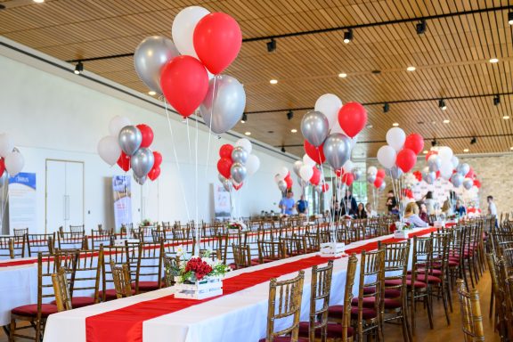 A decorated dining hall with red, white, and silver balloons and a long table setting.