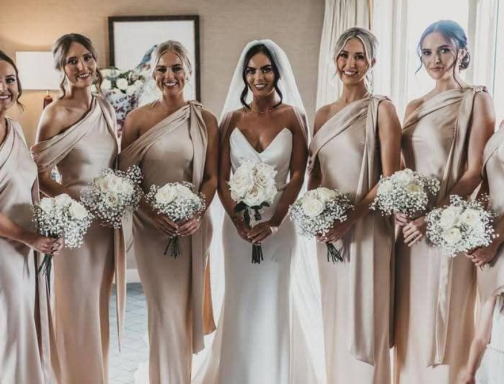 A bride in a white gown stands with six bridesmaids in beige dresses, all holding bouquets.