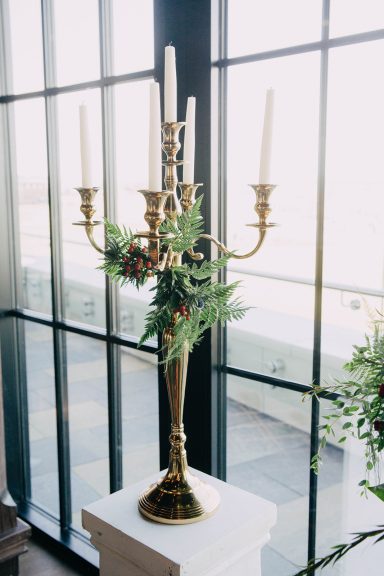 A gold candelabrum with candles and greenery, placed near large windows.
