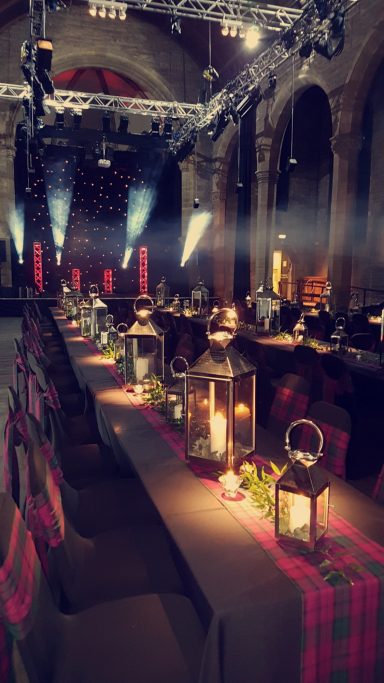 Elegant dining setup with lanterns and candles in a grand hall, softly illuminated.