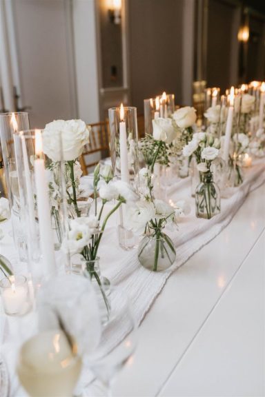 Elegant table setting with white flowers, candles, and glass vases on a white tablecloth.